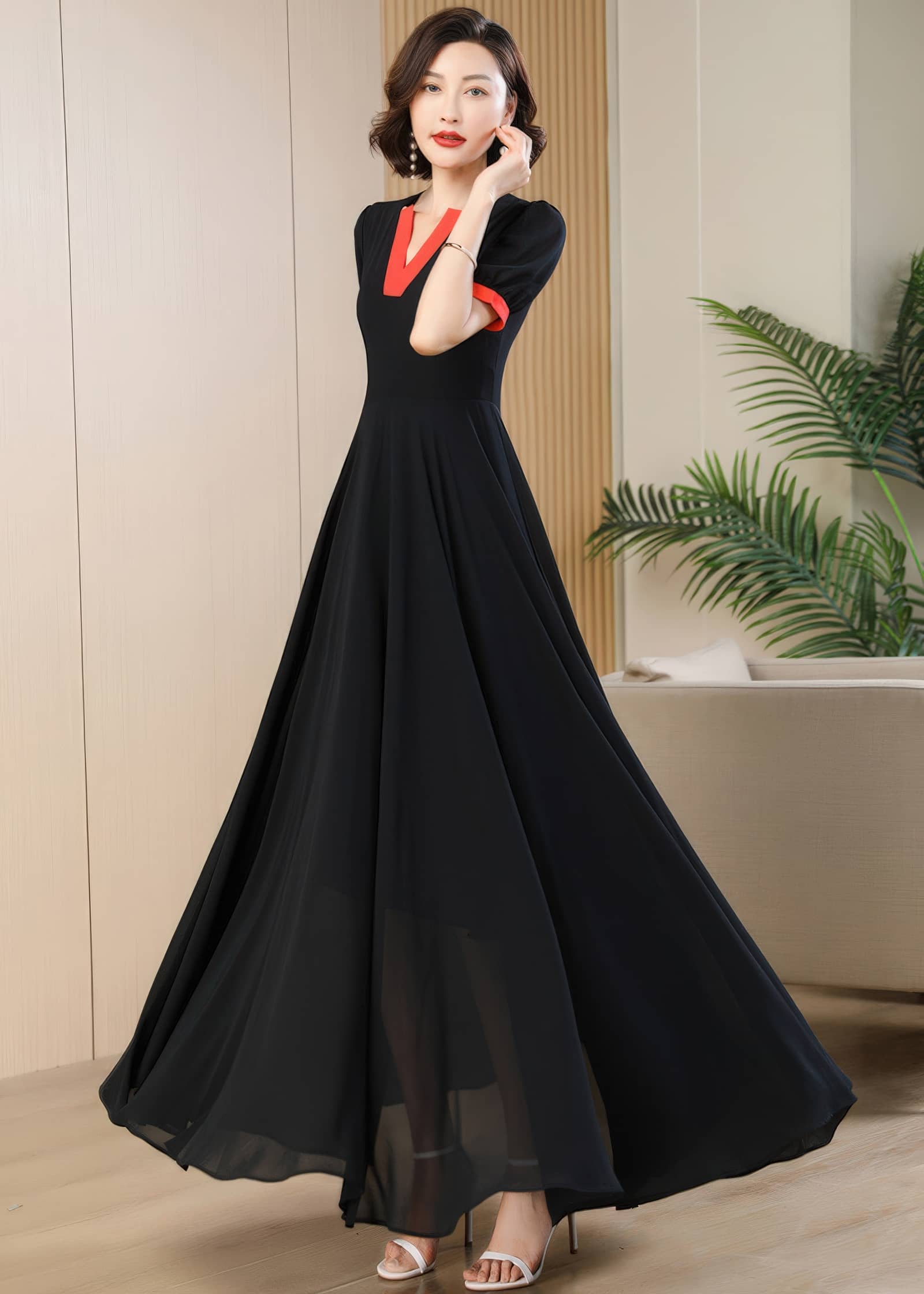 The woman poses mid-stride, showcasing the full flare of the black chiffon A-line maxi dress. Her hand is raised slightly to her face, and the dramatic sweep of the wide, floor-length skirt is emphasized as it catches the air.