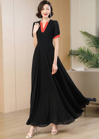 A full-length portrait of the woman standing in the modern black dress with orange-red contrast details. The dress features a high-waisted, fitted bodice that flows into a voluminous chiffon maxi skirt, paired with high-heeled white sandals.