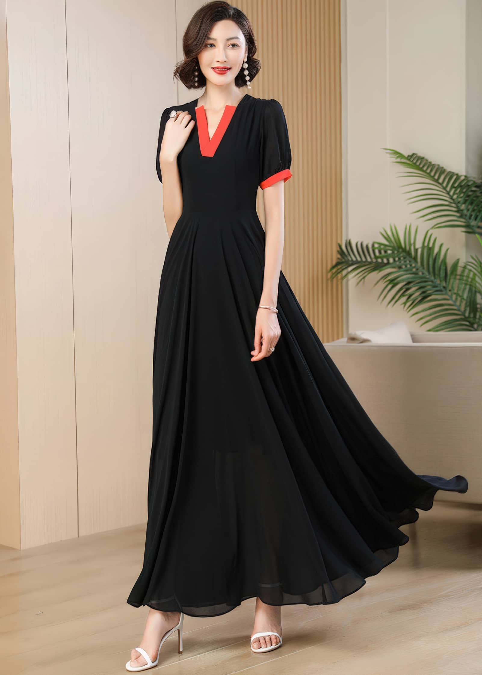 A full-length portrait of the woman standing in the modern black dress with orange-red contrast details. The dress features a high-waisted, fitted bodice that flows into a voluminous chiffon maxi skirt, paired with high-heeled white sandals.