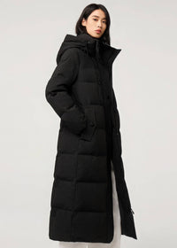A woman wearing a long black hooded puffer coat with a subtle grid texture, photographed against a plain light gray studio background.