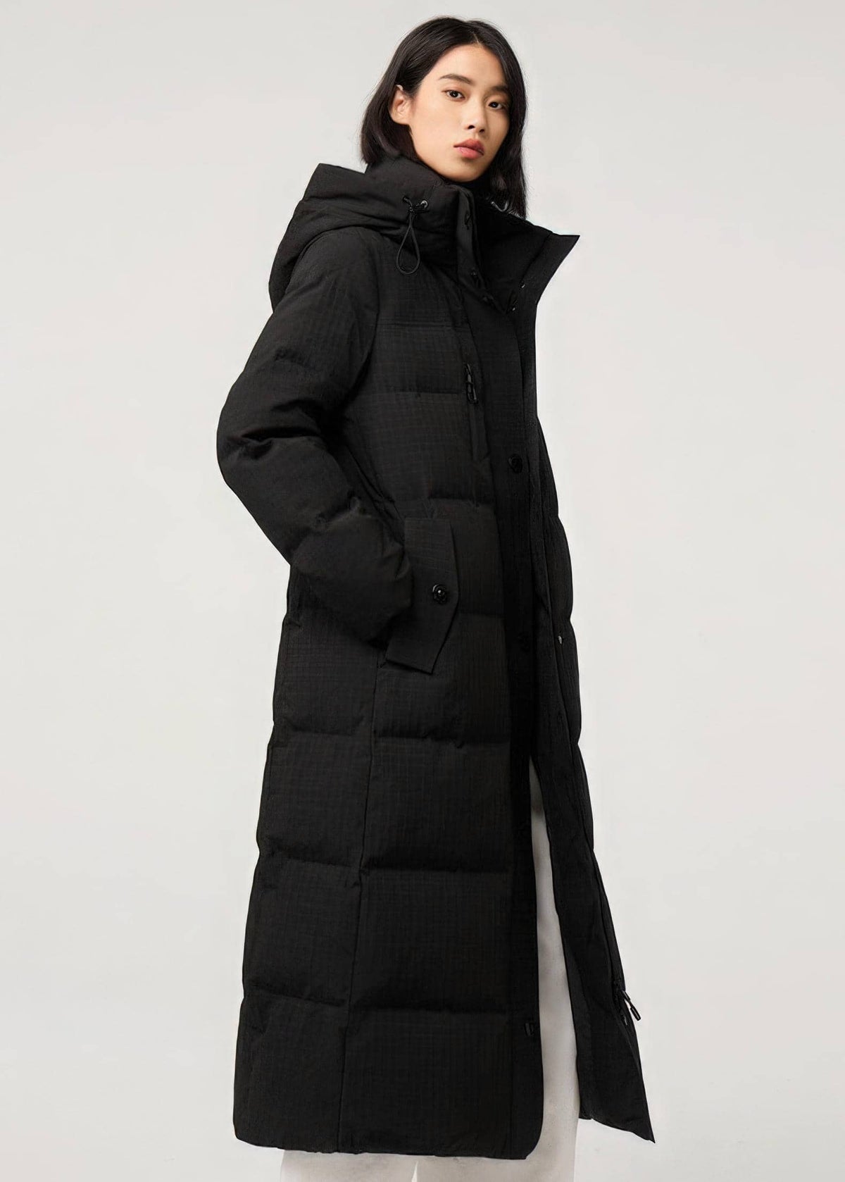 A woman wearing a long black hooded puffer coat with a subtle grid texture, photographed against a plain light gray studio background.