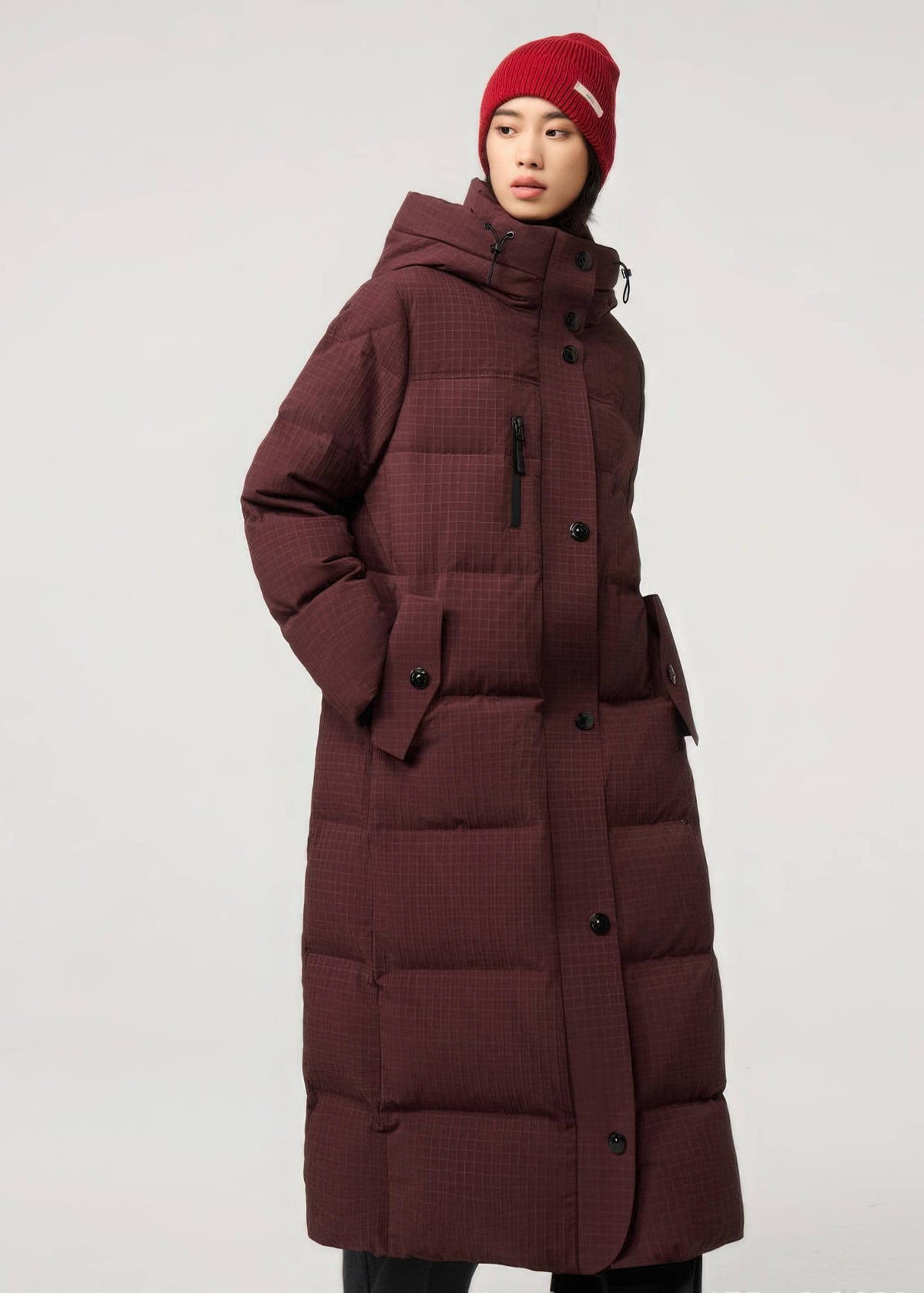 A model wearing a long, dark burgundy puffer coat with a subtle grid pattern, black buttons, and a matching red knit beanie against a light gray studio background.