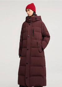 A model wearing a long, dark burgundy quilted puffer coat with a high collar and black buttons, paired with a red knit beanie, against a plain light gray studio background.