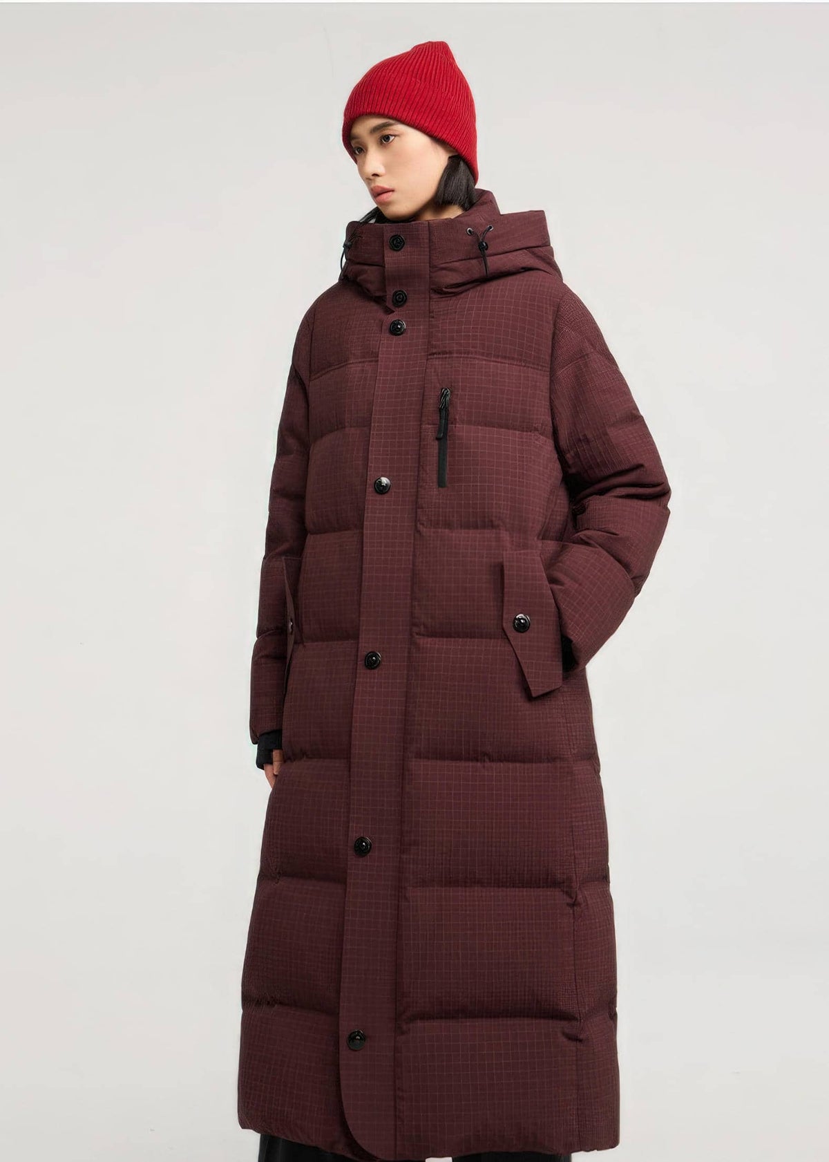 A model wearing a long, dark burgundy quilted puffer coat with a high collar and black buttons, paired with a red knit beanie, against a plain light gray studio background.