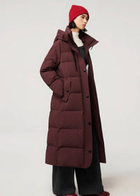 A model wearing a long, dark burgundy quilted puffer coat with a subtle grid pattern, paired with black wide-leg pants, a white turtleneck, and a red knit beanie, set against a plain light gray studio background.