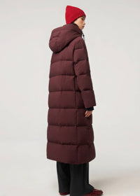 A model shown from the back wearing a long, dark burgundy quilted puffer coat, a red knit beanie, black wide-leg pants, and red shoes, set against a plain light gray studio background.
