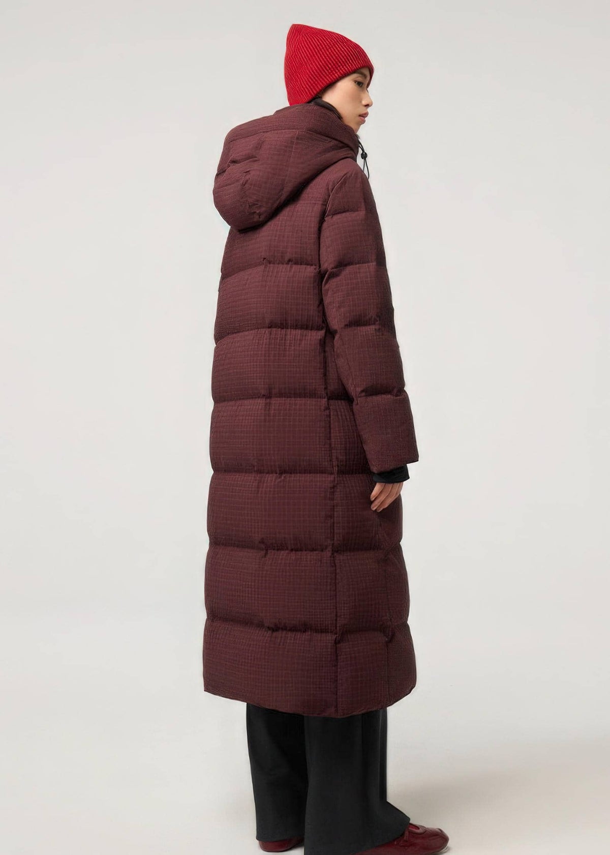 A model shown from the back wearing a long, dark burgundy quilted puffer coat, a red knit beanie, black wide-leg pants, and red shoes, set against a plain light gray studio background.