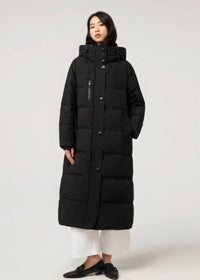 A model wearing a long black quilted puffer coat with a high collar, front zipper and button closure, paired with white cropped pants and black loafers, shot against a plain light gray studio background.