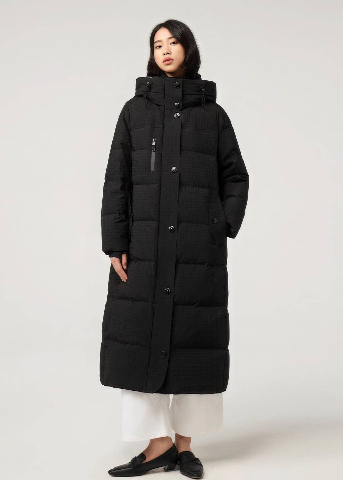 A model wearing a long black quilted puffer coat with a high collar, front zipper and button closure, paired with white cropped pants and black loafers, shot against a plain light gray studio background.
