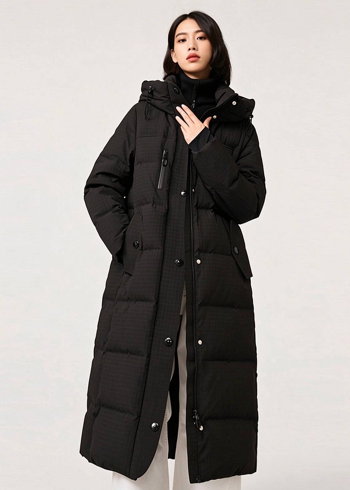A woman modeling a long black quilted puffer coat with a hood, front zipper and snap closure, and large front pockets, photographed against a plain light gray studio background.