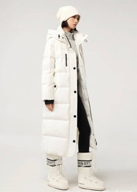 A model wearing a long white puffer coat with a hood, black buttons, and black zipper details, styled with a matching white beanie, gray sweater, black pants, and white snow boots against a light gray studio background.
