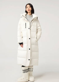 A model wearing a long white quilted puffer coat with black buttons and a high collar, paired with a gray turtleneck, white pants, and white boots with black lettering, set against a plain light gray studio background.