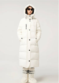 A model wearing a long white puffer coat with a hood, black snap buttons, and a vertical ribbed texture, paired with white snow boots and patterned socks, against a light gray studio background.