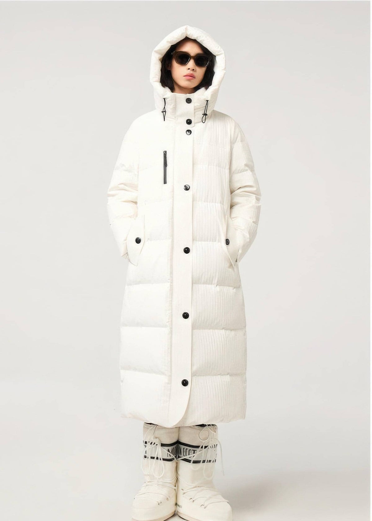 A model wearing a long white puffer coat with a hood, black snap buttons, and a vertical ribbed texture, paired with white snow boots and patterned socks, against a light gray studio background.