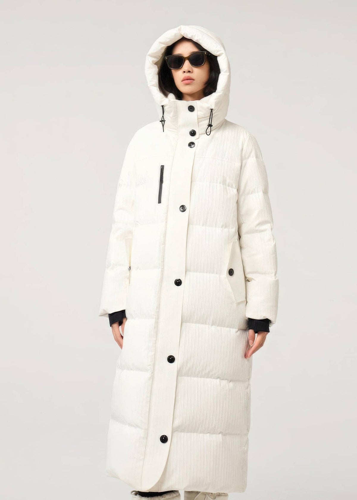 A model wearing a long, hooded puffer coat in off-white with black buttons and black ribbed cuffs, set against a plain light gray studio background.