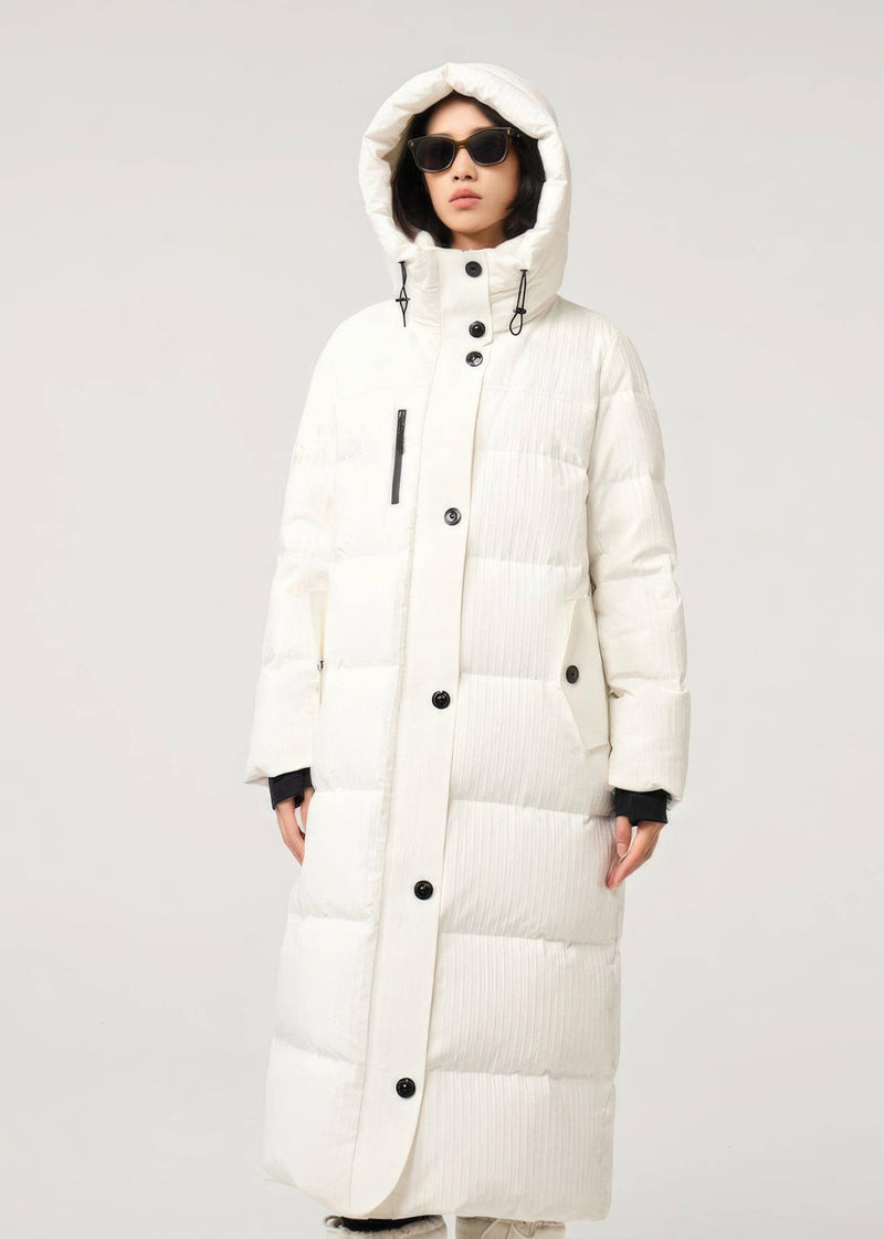 A model wearing a long, hooded puffer coat in off-white with black buttons and black ribbed cuffs, set against a plain light gray studio background.