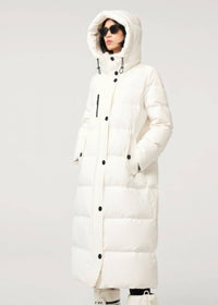 A model wearing a long, hooded puffer coat in off-white. The coat features horizontal quilting, black snap buttons, and black drawstrings.