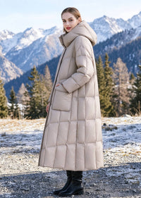 Side profile view of woman wearing beige longline down parka with flap pockets against snow mountain background