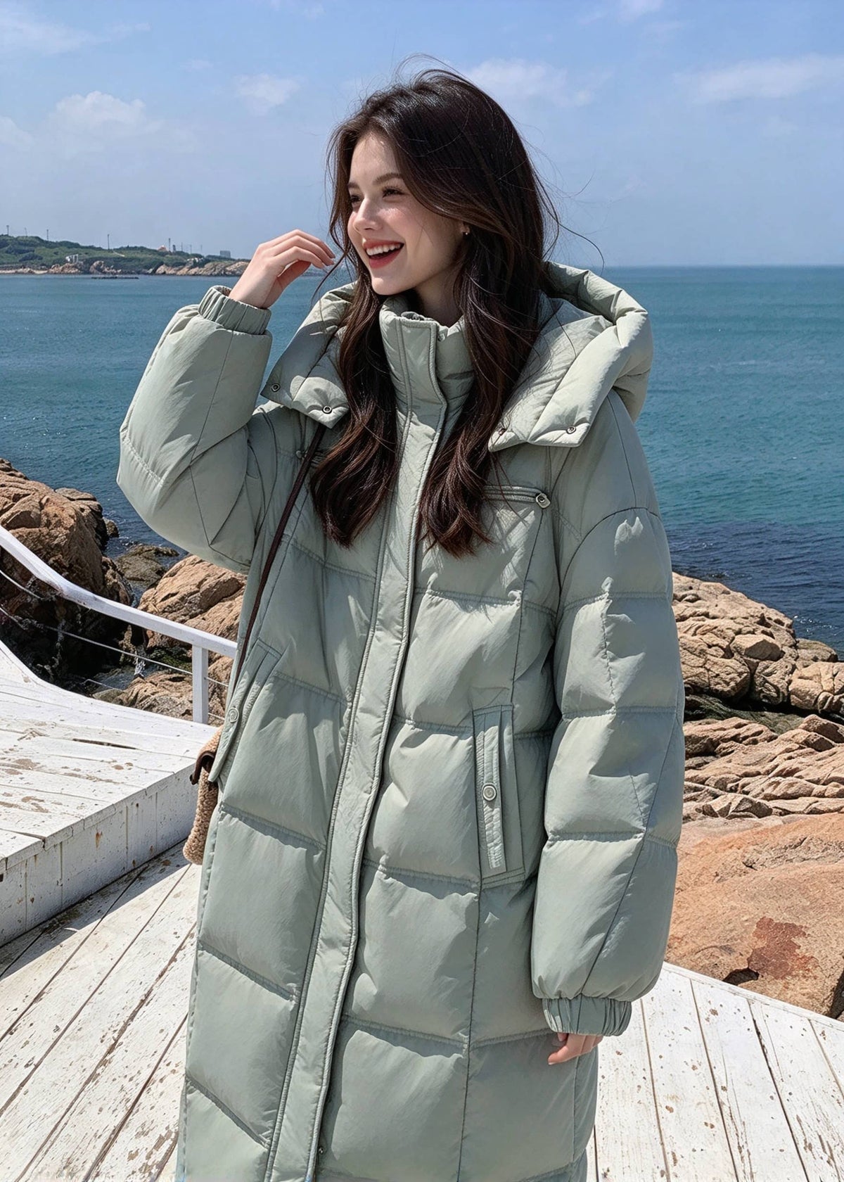 Woman smiling and touching hair wearing sage green quilted hooded goose down long coat