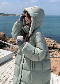 Side view of woman with hood up holding coffee cup wearing sage green high stand collar goose down long coat