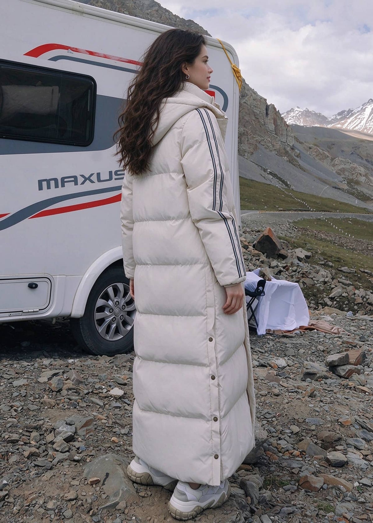 Full length back profile view of woman wearing off-white side-stripe longline puffer coat with detachable hood near snowy mountains