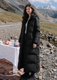 Full length front view of smiling woman wearing black side-stripe longline puffer coat with detachable hood near outdoor campsite