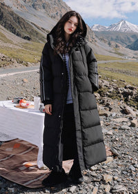 Full length front portrait of woman wearing black side-stripe longline puffer coat with hood against mountain backdrop