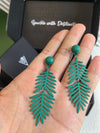 A person holding a pair of the matte green, fern-leaf dangle earrings in their open palm, providing a scale reference for the jewelry.