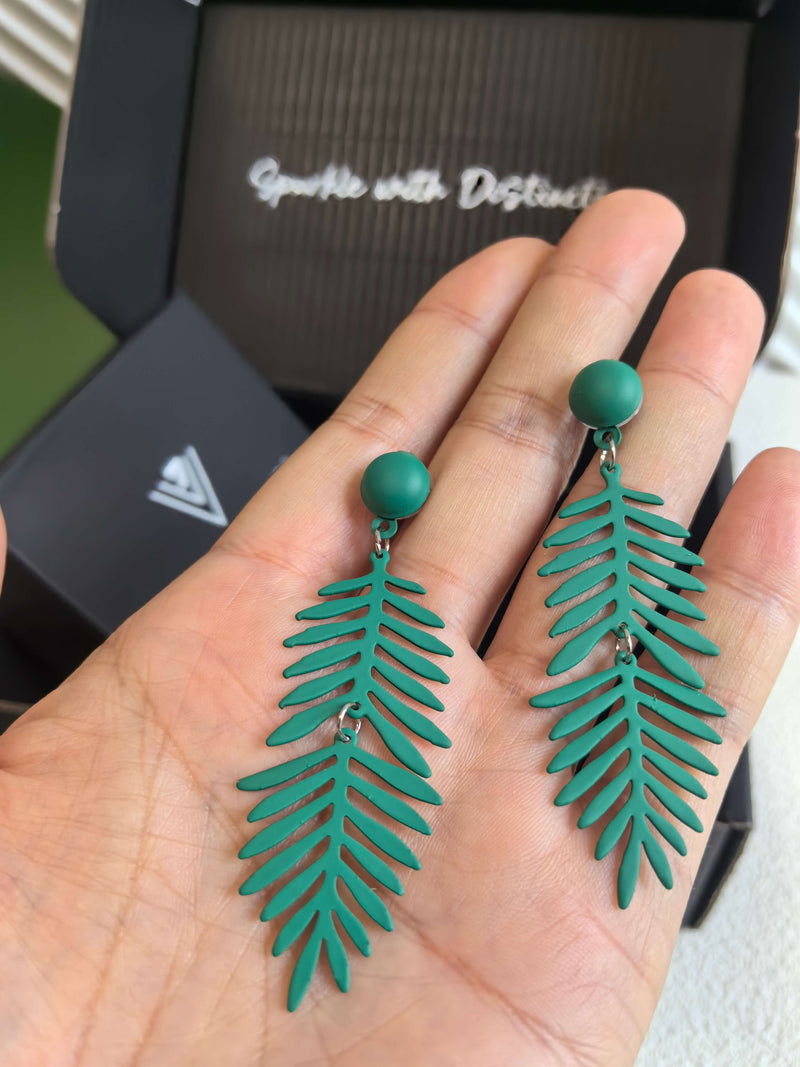 A person holding a pair of the matte green, fern-leaf dangle earrings in their open palm, providing a scale reference for the jewelry.