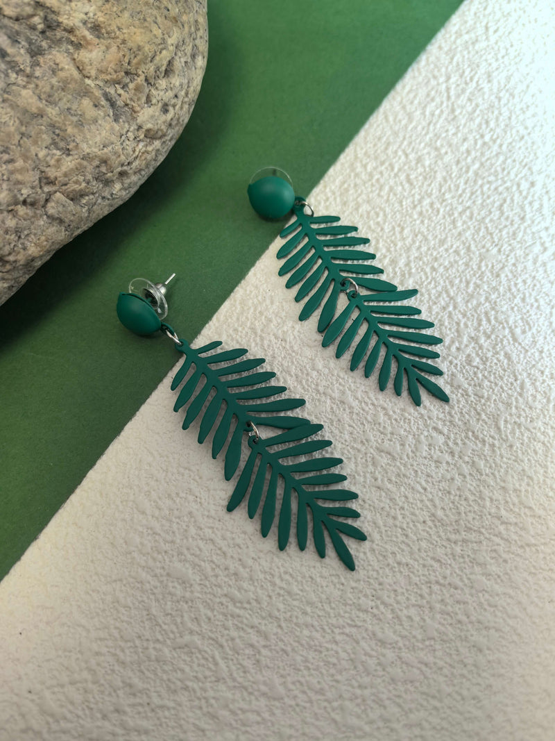 A pair of forest-green, tropical fern-leaf dangle earrings displayed next to a large grey rock on a split green and textured white background.