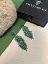A pair of dark green fern-leaf earrings displayed next to a closed black jewelry box labeled ""VIVIAN SEVEN,"" on a background split between green and white.