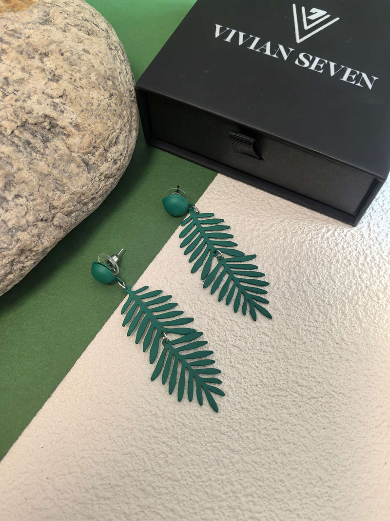A pair of dark green fern-leaf earrings displayed next to a closed black jewelry box labeled ""VIVIAN SEVEN,"" on a background split between green and white.