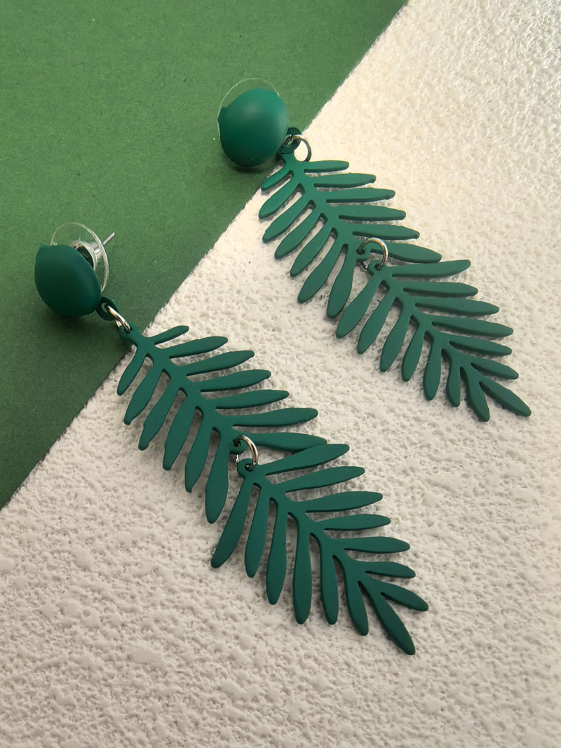 A pair of long, forest-green fern-leaf dangle earrings lying on a backdrop split between dark green and textured white. The earrings feature a tiny jump ring in the middle of the leaf section.