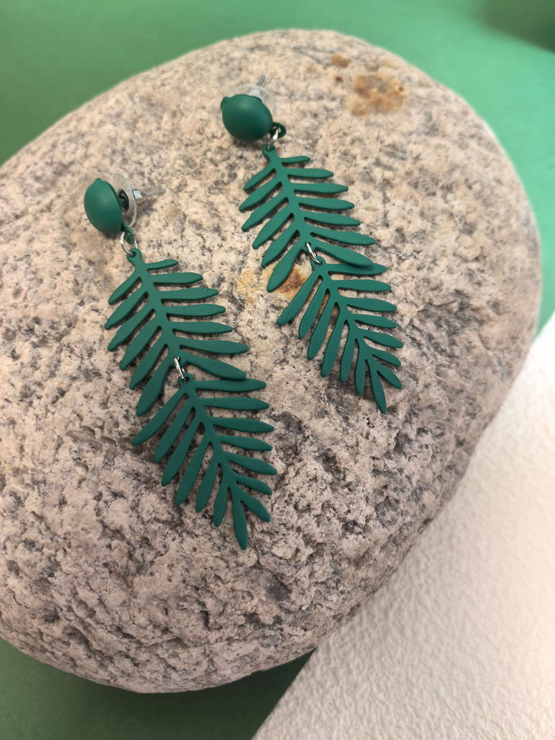 A pair of matte green fern-leaf dangle earrings displayed on a large, rough-textured grey rock.