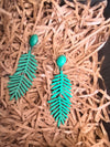 A pair of matte forest-green, dangle earrings shaped like fern leaves, resting on a bed of shredded, light-brown packing paper.