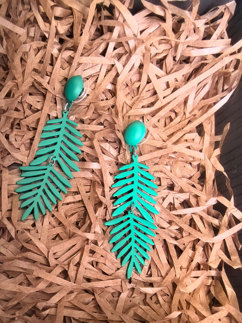 A pair of matte forest-green, dangle earrings shaped like fern leaves, resting on a bed of shredded, light-brown packing paper.