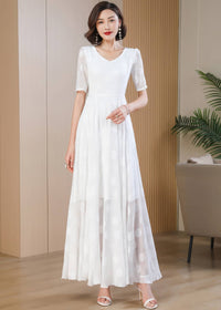 A full-length front view of a woman standing and modeling a white maxi dress with large, subtle, tonal polka dots. The dress features a V-neckline, short sheer sleeves, and a flowing A-line skirt with a sheer hem overlay. She is wearing white strappy high heels and pearl drop earrings.