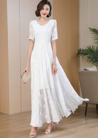 A full-length shot of a woman walking forward, showcasing the movement of a white polka-dot maxi dress. She holds a small gold clutch bag in her right hand, wearing white strappy heels and elegant pearl earrings.