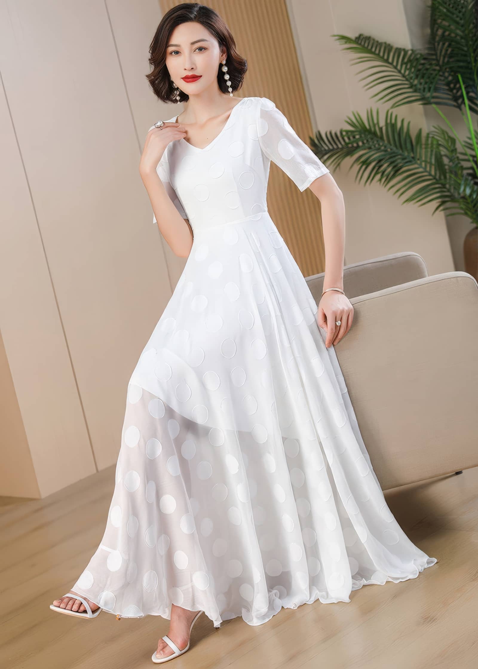A full-length image of a woman leaning elegantly against the back of a beige armchair. She is wearing a white V-neck maxi dress with a faint polka-dot pattern, white high-heeled sandals, and large pearl drop earrings.
