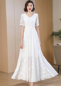 A front-facing, full-length photo of a woman standing as the skirt of her white polka-dot maxi dress flares out slightly at the sides. She is styled with short, wavy dark hair, red lipstick, pearl earrings, and white high heels.