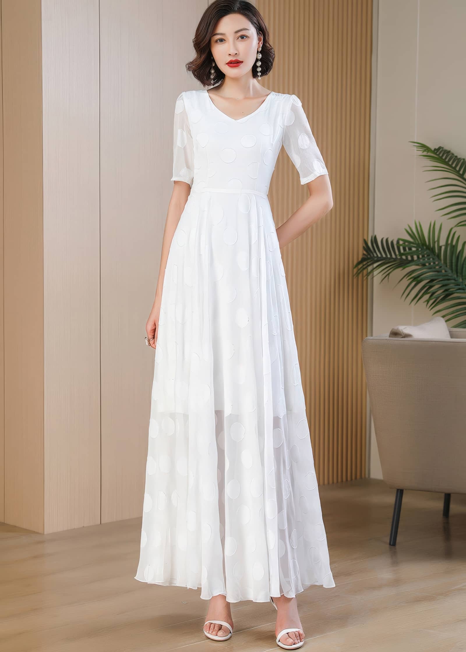 A full-length front view of a woman posing with one hand behind her back, wearing a white short-sleeved maxi dress with large, tonal polka dots. The sheer bottom portion of the skirt reveals the silhouette of her legs.