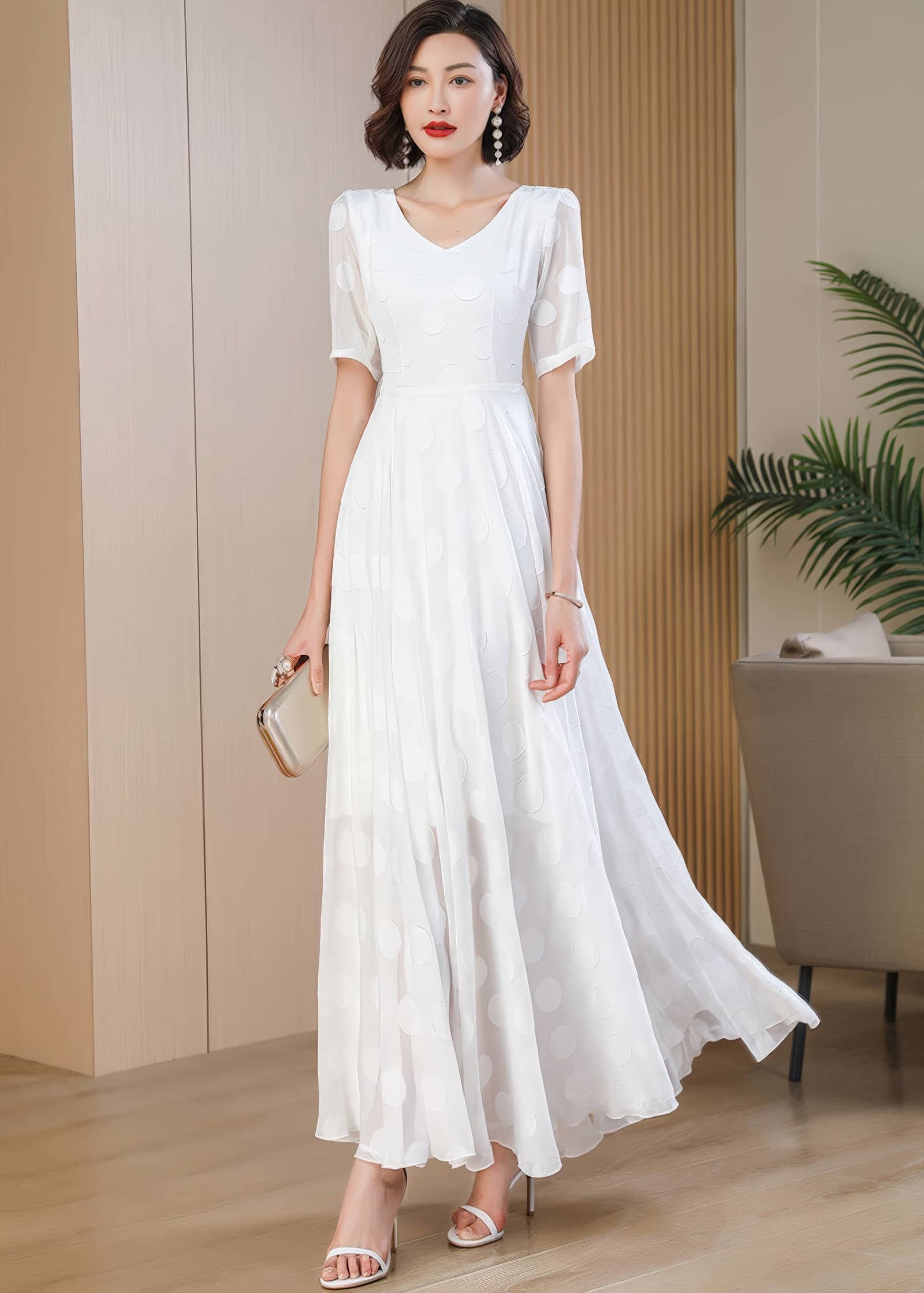 A full-length shot of a woman in motion, mid-stride, carrying a gold evening clutch. She wears a white polka-dot maxi dress with a flowing skirt, paired with white strappy sandals.