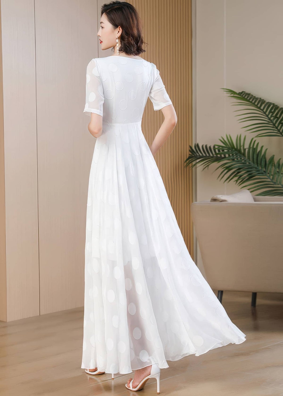 A full-length side and back profile view of a woman looking over her shoulder, displaying the back design of a white polka-dot maxi dress. The fabric is light and flowing, showing the movement of the skirt as she steps forward in white stiletto sandals.