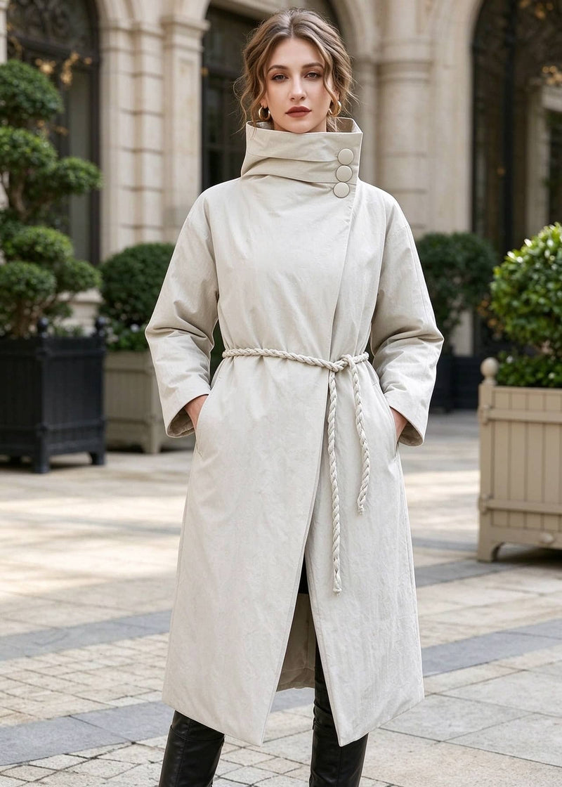 Woman with hands in pockets wearing beige asymmetric high neck longline down coat with braided rope waist belt