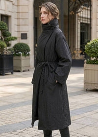 Side view of woman in black high neck longline down coat with side hand pockets and rope waist belt