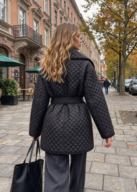 A woman wearing a black quilted belted coat and dark wide-leg trousers stands on a city sidewalk, photographed from the back. The street features historic brick buildings, trees with autumn leaves, and a few pedestrians in the distance.
