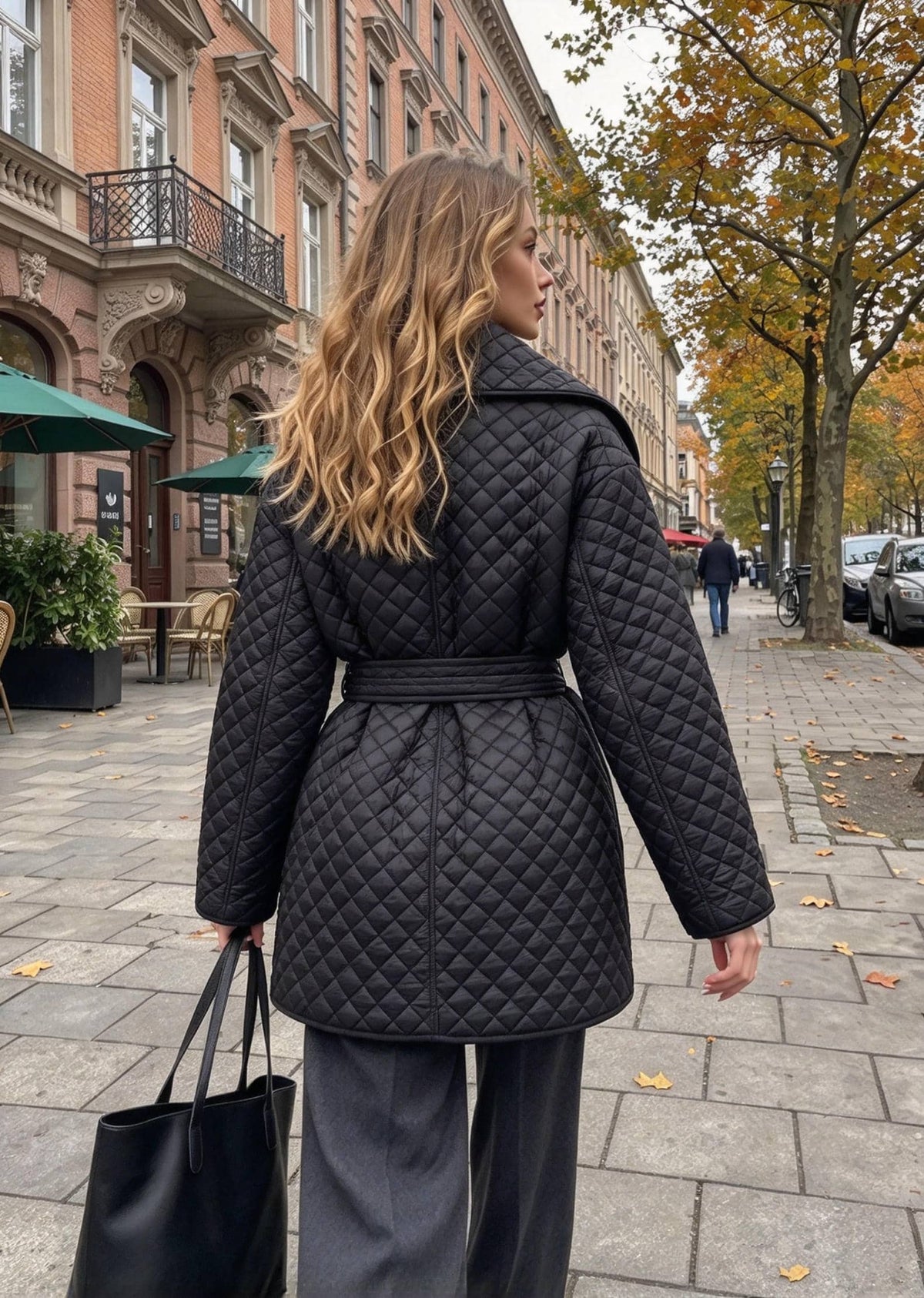 A woman wearing a black quilted belted coat and dark wide-leg trousers stands on a city sidewalk, photographed from the back. The street features historic brick buildings, trees with autumn leaves, and a few pedestrians in the distance.