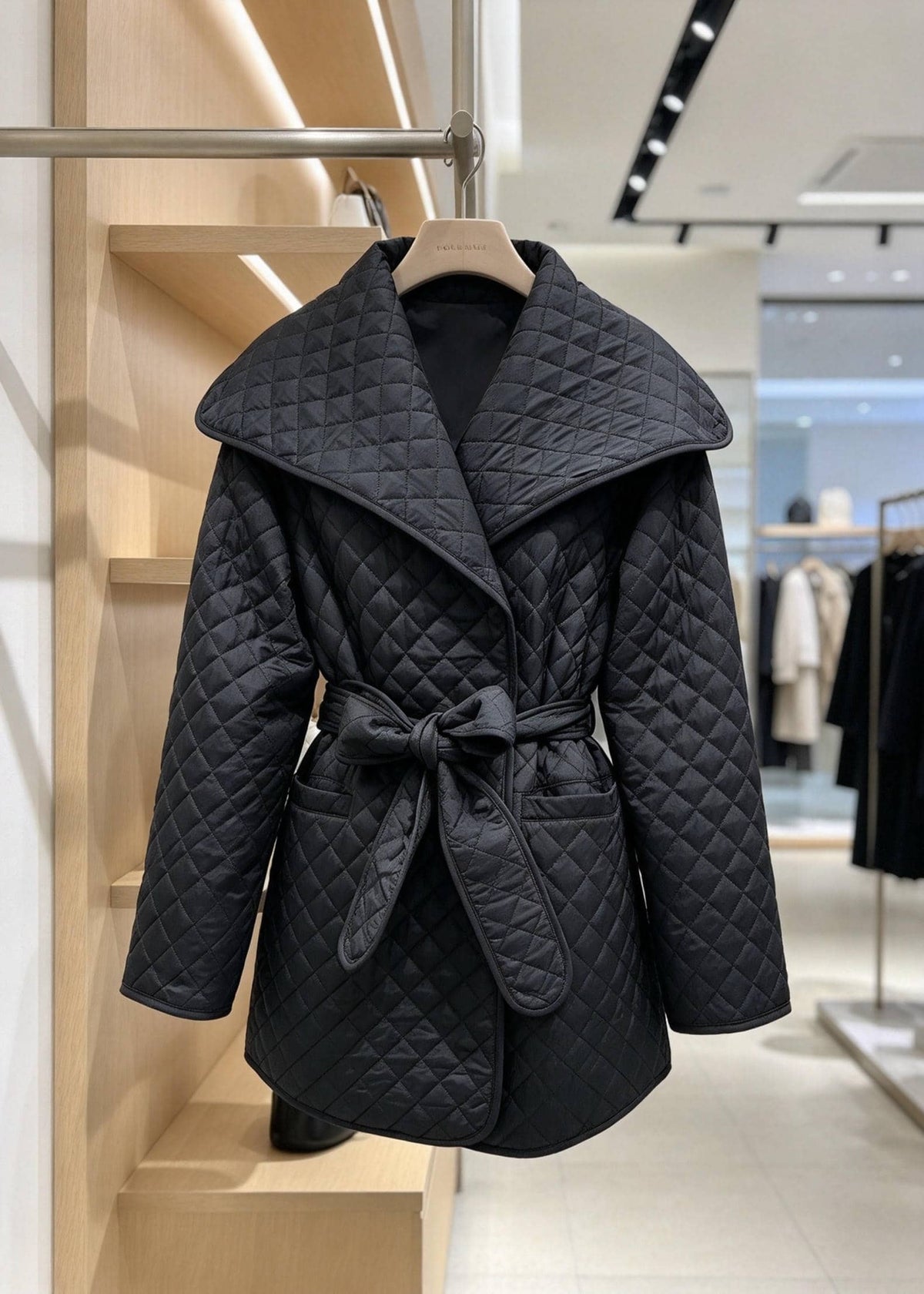 A black quilted wrap coat with a wide collar and self-tie belt, hanging on a wooden hanger in a boutique setting.