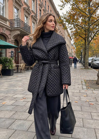 A woman walking on a city sidewalk wearing a black quilted wrap coat over a black turtleneck, dark wide-leg pants, and carrying a black tote bag.