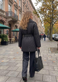 A woman walking on a city sidewalk wearing a black quilted belted coat, dark wide-leg trousers, and carrying a black tote bag.